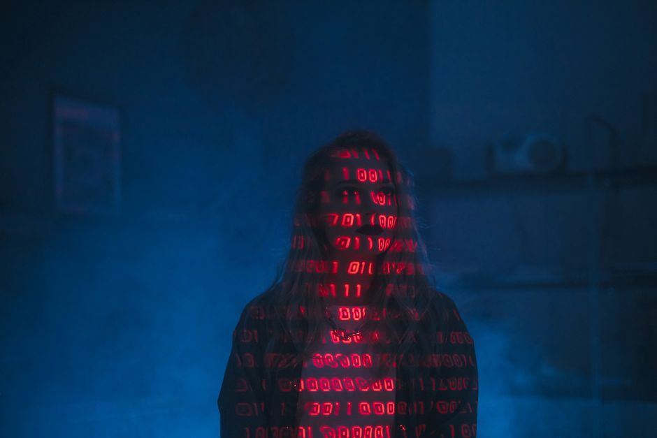 A person wearing a dark hoodie standing in a dimly lit environment with a blue hue, their face partially obscured by a digital display of red binary code projecting onto their face and clothing. The b