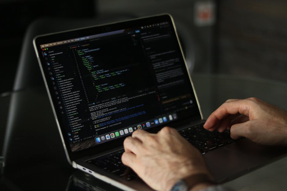 A person wearing a dark hoodie standing in a dimly lit environment with a blue hue, their face partially obscured by a digital display of red binary code projecting onto their face and clothing. The b