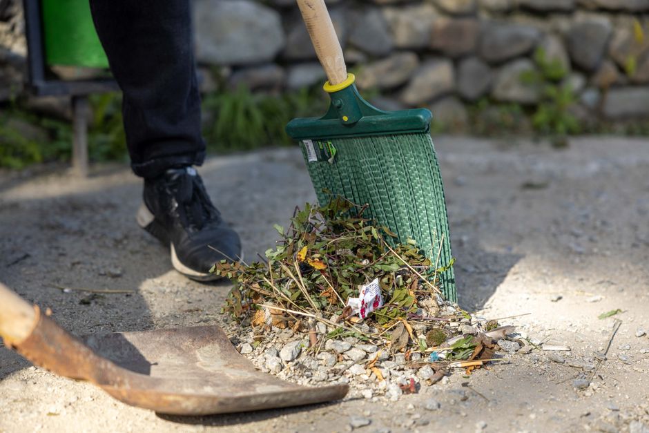 A person wearing black pants and black shoes is raking a small pile of garden debris, including leaves, twigs, and some litter, onto a green garden waste bin with a yellow band near the handle. The bi